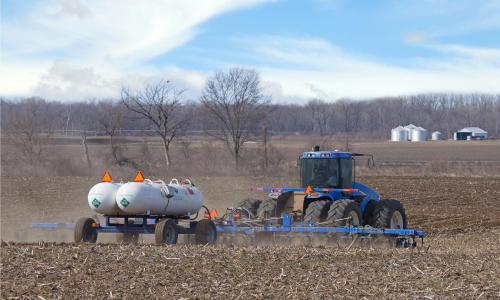 photo of a tractor in a turned-over field pulling a trailer with two tanks of anhydrous ammonia fertilizer that is being sprayed on the field; farm buildings 和 silos are in the distance